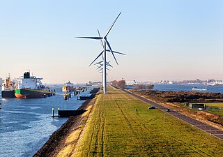 Windkraftanlagen auf einem Deich, Frachtschiffe im Wasser und Hafen von Rotterdam im Hintergrund.